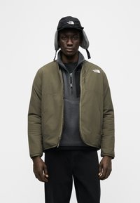 Άνδρας που φορά πράσινο χακί επενδεδυμένο μπουφάν και γκρι φλις με φερμουάρ, με μαύρο παντελόνι και μαύρο χειμερινό καπέλο με καλύμματα αυτιών, στέκεται μπροστά σε λευκό φόντο.