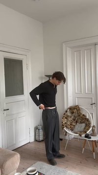 Black long-sleeve shirt, gray pleated trousers with a belt loop, and dark shoes; standing indoors near a chair and decorative items.