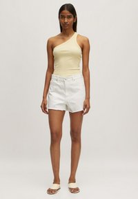 Één-schouder lichtbeige top gecombineerd met hooggetailleerde witte denimshorts met een rafelige onderkant. Zachte textuur, minimalistisch ontwerp, geen zichtbare patronen.