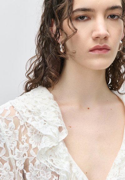 Νεαρή γυναίκα με καστανά σγουρά μαλλιά, που φορά ασημένια σκουλαρίκια κρίκους και λευκό δαντελένιο τοπ με γιακά με βολάν, ουδέτερη έκφραση.