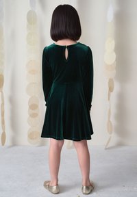 Barn med kort hår iført en mørkegrøn fløjlsnederdel med lange ærmer og guld sko, stående med ryggen til kameraet på en lys gulv.