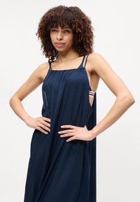 Γυναίκα με σγουρά μαλλιά φορώντας ένα φαρδύ μπλε σκούρο αμάνικο φόρεμα, στέκεται με τα χέρια στη μέση μπροστά από λευκό φόντο.