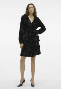 Robe portefeuille noire en tissu pelucheux avec des sequins, dotée de manches longues, d'une taille à nouer et d'une jupe évasée. Associée à des talons hauts noirs.