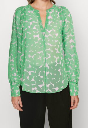 Kvinde iført en grøn bluse med lange ærmer med et lyst rosa abstrakt mønster og V-hals, kombineret med sorte bukser.