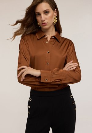 Bruine satijnen shirt met een kraag en knoopsluiting, gecombineerd met hoge zwarte broeken met gouden oogjes accenten.