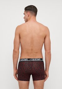 Boxer breve nero con un motivo di ciliegie rosse, dotato di una vita spessa con l'etichetta "Y&SONS". Il tessuto sembra liscio e aderente.