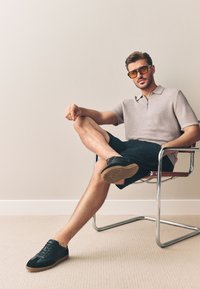 Polo gris texturé avec une demi-fermeture éclair, short noir et baskets noires avec semelles en caoutchouc. Modèle assis sur une chaise chromée.