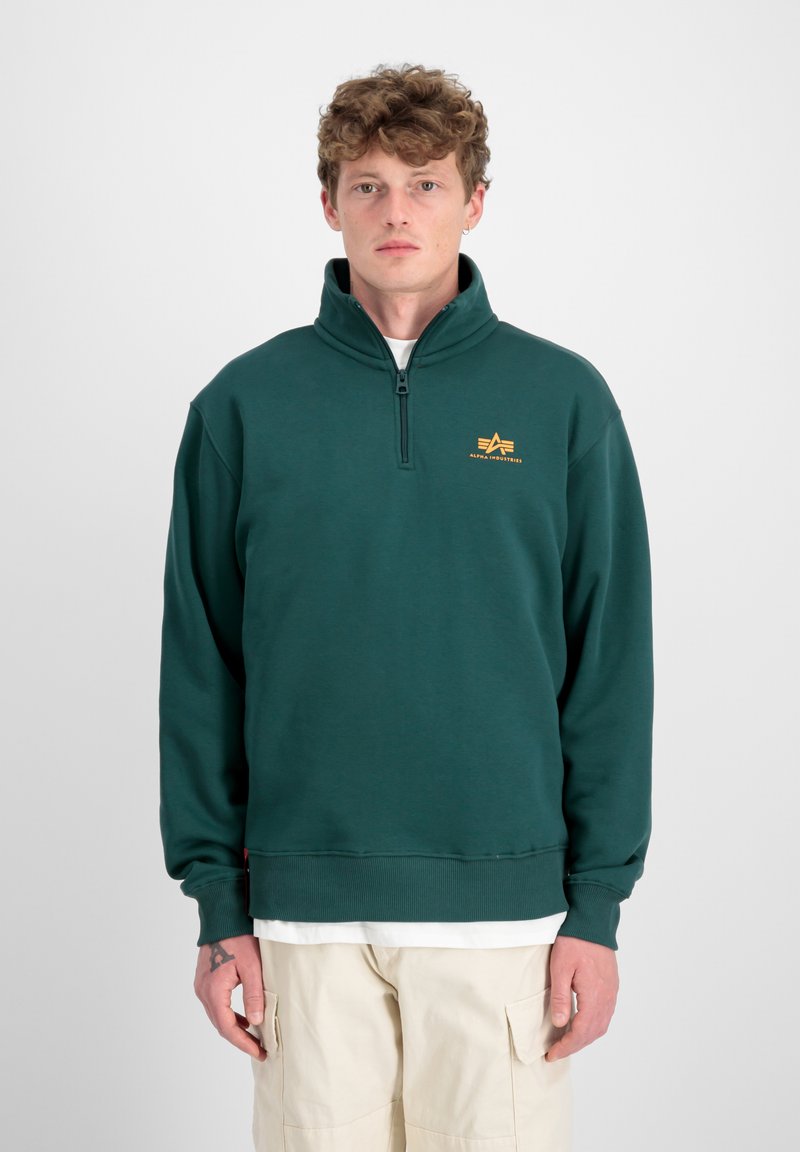 Uomo che indossa un felpa verde scuro con zip a quarto e logo giallo, e pantaloni cargo beige, in piedi su uno sfondo neutro.