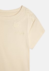 T-shirt a costine crema con ricami floreali sulla spalla e il nome del marchio ricamato in tono sul davanti.