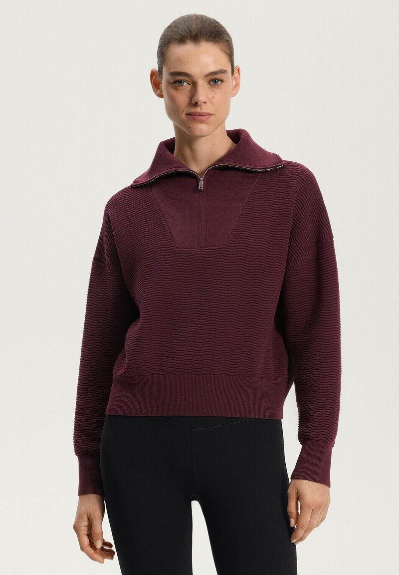 Pulover cu fermoar burgundy, cu textură de tip ribbing, guler larg și croi lejer, asociat cu colantii negri, pe un fundal neutru.