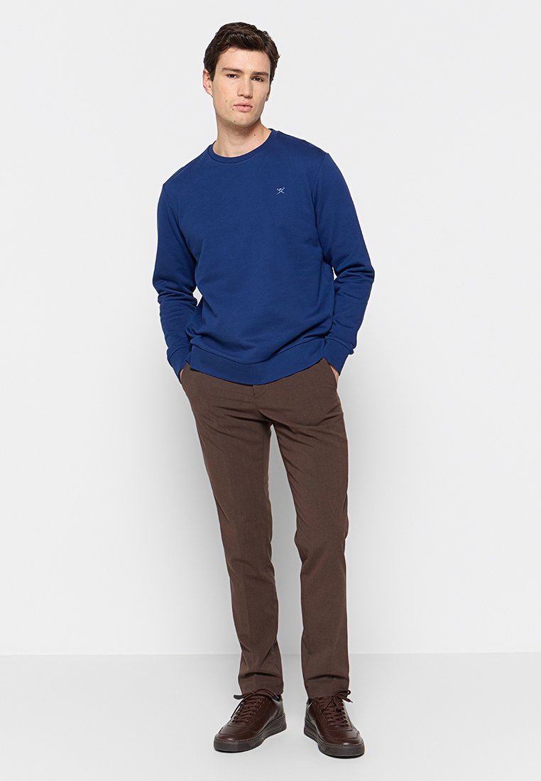 Sudadera azul de algodón, con cuello redondo y puños acanalados, combinada con pantalones marrones y zapatillas oscuras.