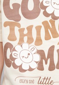 T-shirt crema con grande testo marrone che recita "GO THINGS" e una grafica di un fiore sorridente con una scintilla, contornata di marrone.