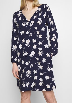 Femme portant une robe bleu marine à col en V avec manches longues et motif floral blanc, debout devant un fond clair uni.