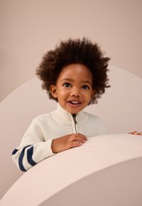 Maglione bianco a coste con zip e dettagli a righe blu sulle maniche, indossato da un bambino con i capelli ricci, che sorride su uno sfondo morbido.
