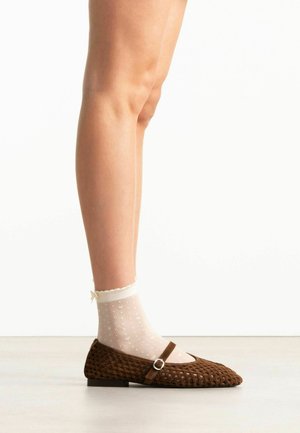 Been met doorschijnende witte enkelsokken met kleine hartjespatronen en een bruine geweven Mary Jane platte schoen met gesp.