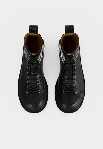 Ζευγάρι μαύρες δερμάτινες μπότες με κορδόνια, με διάτρητο σχέδιο στα δάχτυλα και ταινίες τραβήγματος στη φτέρνα, εμφανιζόμενες από πάνω σε λευκό φόντο.