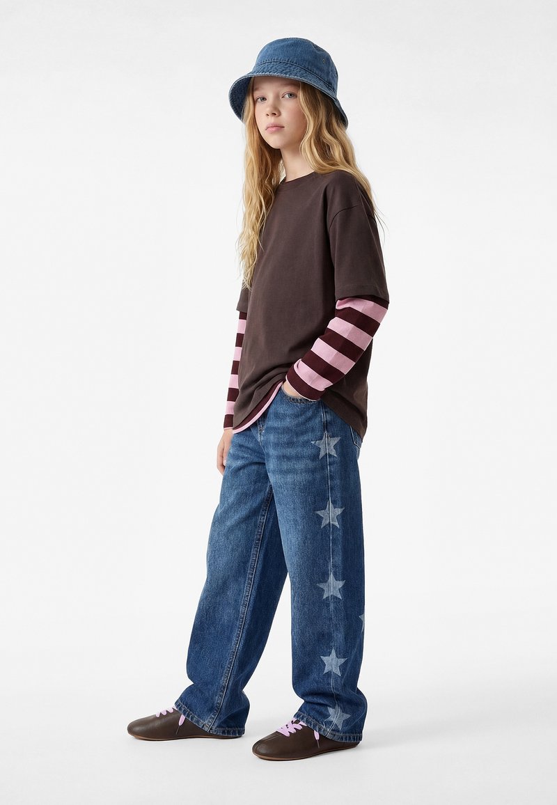 Giovane che indossa un cappello a secchiello di denim blu, una maglietta marrone sopra una camicia a maniche lunghe a righe rosa, jeans blu con stampe di stelle e scarpe marroni.