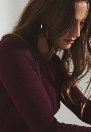 Bordeauxfarbene Bluse mit langen Ärmeln und taillierter Passform aus einem strukturierten Strickmaterial. Das Haar fällt sanft über die Schulter und betont den Stoff.