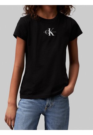 Chica con camiseta negra de Calvin Klein Jeans y vaqueros azules de mezclilla, posando con una mano en el bolsillo frente a un fondo gris liso.