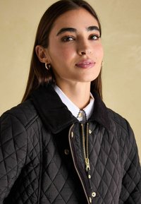 Gep quiltete schwarze Jacke mit einem Cordkragen, goldenen Reißverschluss und Druckknöpfen. Getragen über einem weißen Hemd mit Kragen und dekorativen Nähten.