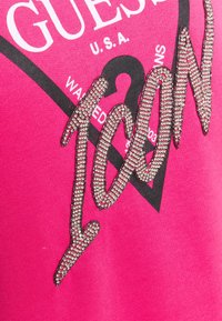 Růžová látka s velkým černým textovým grafikem "GUESS U.S.A." a dekorativními stříbrnými kamínky tvořícími slovo "ICON."