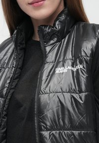 Κοντινό πλάνο προσώπου που φοράει μαύρο γυαλιστερό μπουφάν χωρίς μανίκια με το λογότυπο "KARL LAGERFELD JEANS" και μαύρο πουλόβερ από κάτω.