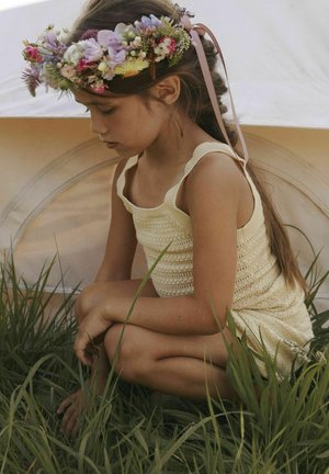 Un bambino è accovacciato nell'erba, indossando un vestito a canotta di maglia gialla chiara. Una corona floreale decorata con fiori rosa e viola poggia sulla sua testa.