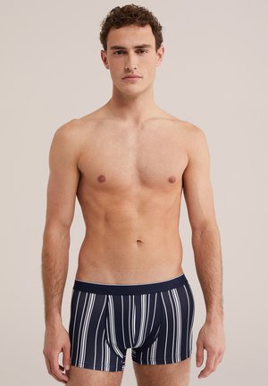 Muž bez trička s kučeravými vlasmi, ktorý má na sebe tmavomodré boxerky s bielymi zvislými pruhmi, stojaci pred jednofarebným svetlým pozadím.