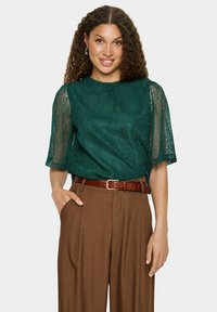 Blouse en dentelle bleu-vert avec des manches courtes et amples, associée à un pantalon marron taille haute et une ceinture marron, présentant une finition texturée.