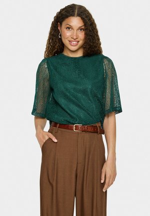 Blouse en dentelle bleu-vert avec des manches courtes et amples, associée à un pantalon marron taille haute et une ceinture marron, présentant une finition texturée.