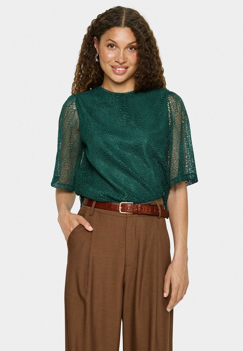 Blouse en dentelle bleu-vert avec des manches courtes et amples, associée à un pantalon marron taille haute et une ceinture marron, présentant une finition texturée.