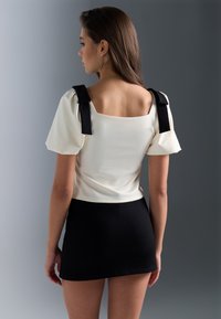 Top blanc avec des accents en ruban noir, dos carré, manches bouffantes et jupe noire ajustée. Texture lisse avec des détails de couleur contrastante.