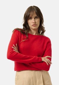 Maglione rosso lavorato a maglia con maniche lunghe e scollatura ampia, esposto su uno sfondo neutro. Texture morbida e vestibilità rilassata.