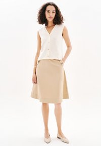 Cremefärgad ärmlös topp med V-ringning och knapprad, matchad med en beige A-line kjol med dekorativa knappar på fickorna.