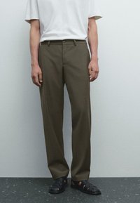 Man som bär olivgröna raka byxor, svarta slutna sandaler och en vit kortärmad T-shirt står mot en enfärgad vägg.