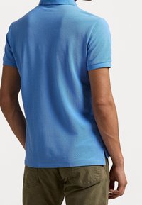 Man in een lichtblauw poloshirt met korte mouwen en olijfgroene broek, van achteren afgebeeld tegen een effen achtergrond.