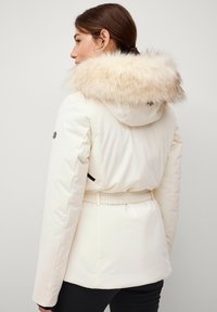 Witte pufferjas met een met bont gevoerde capuchon, getailleerde taille en zwarte accenten. Gladde textuur, lange mouwen en een elastische band in de taille.