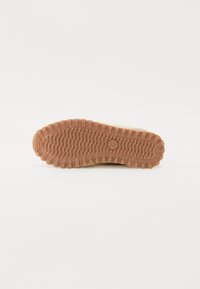 Sål av en beige sko med en teksturert gummioverflate, med et mønstrete dekk og et sirkulært merke i midten. Fargen er lys brun.