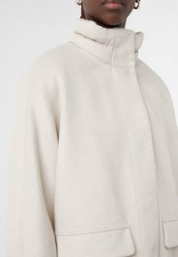 Cappotto in misto lana beige chiaro con colletto alto e imbottito. Presenta una vestibilità rilassata e tasche laterali, enfatizzando un'esterno morbido e testurizzato.