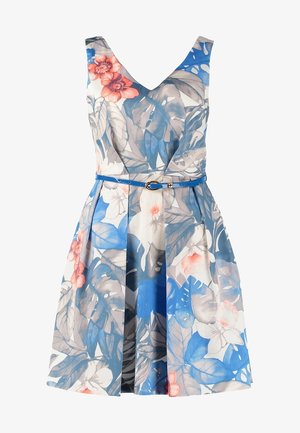 Mujer con vestido sin mangas azul y blanco con estampado floral, cinturón fino azul, sujetando gafas de sol, caminando con zapatos blancos con cordones.