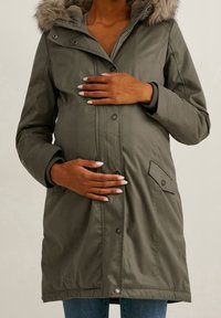 Oliwkowo zielona parkę ciążowa z kapturem wyłożonym futrem, przednimi kieszeniami i zamkiem błyskawicznym. Luźny krój odpowiedni dla przyszłych mam.