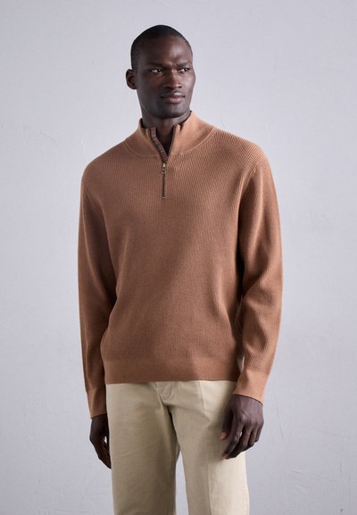 Brauner gerippter Pullover mit einem Viertelreißverschluss und einem bunten Innenkragen. Kombiniert mit hellbeigen Hosen, vor einem neutralen Hintergrund präsentiert.