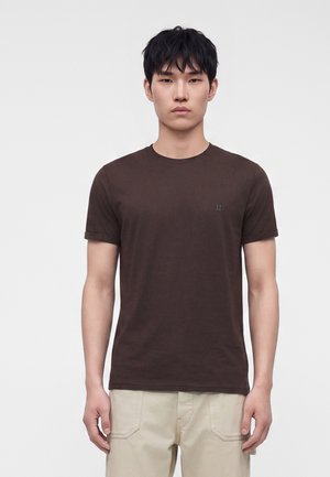 Mand med kort sort hår iført en ensfarvet mørkebrun T-shirt og lyse beige bukser står foran en hvid baggrund.