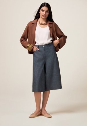 Bruine korte mouwenjacket over een witte top, gecombineerd met denim culottes. Accessoires zijn een chunky gouden ketting en armbanden, en beige ballerina's.