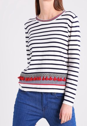 Donna che indossa una camicia bianca a manica lunga con righe blu navy e nappine rosse lungo l’orlo, abbinata a jeans blu.