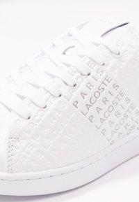 Zapatilla blanca con patrón de texto en relieve, con cordones y una superficie texturizada. La suela es lisa con un diseño minimalista.