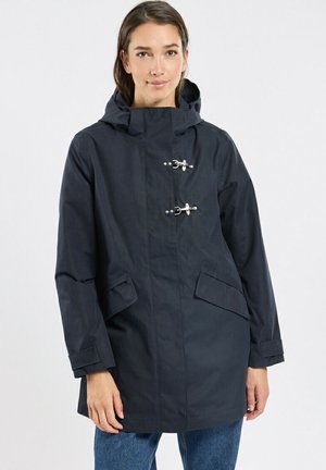 Femme portant un imperméable à capuche bleu marine avec deux fermetures à bascule en métal et des poches à rabat sur le devant, debout devant un fond clair uni.