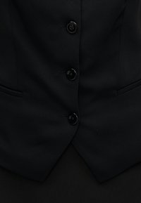 Μαύρο ραμμένο γιλέκο με απαλό ύφασμα, έξι στρογγυλά κουμπιά και δύο πλαϊνές τσέπες, με κομψό γιακά με κορυφή σε αιχμηρό σχήμα.