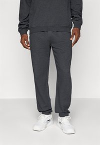 Mann iført mørkegrå joggebukser og matchende hettegenser, kombinert med hvite joggesko, som står mot en lys, ensfarget bakgrunn.