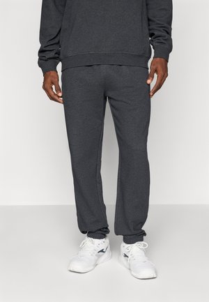 Homme portant un pantalon de survêtement gris foncé et un sweat-shirt assorti, associé à des chaussures de sport blanches, se tenant devant un fond clair.